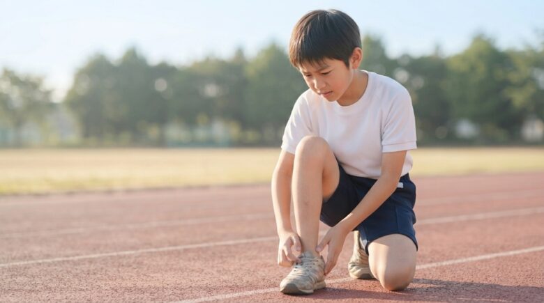 陸上のトラックで練習中にかかとの痛みを気にしてしゃがみ込む小学生の男の子のイメージ