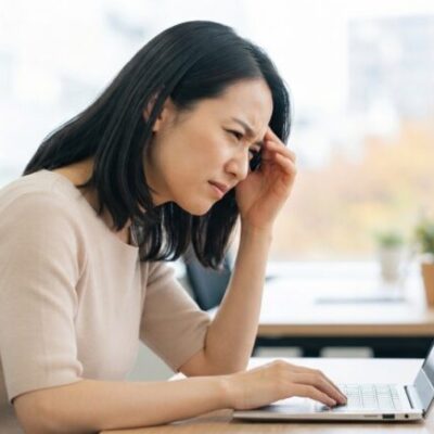 デスクでノートパソコンをのぞき込み、こめかみに手を当てて頭痛をこらえる女性