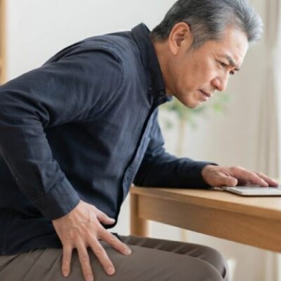デスクワークで背中が丸くなり顔が前に出た姿勢のまま、太ももに手を当てる男性(イメージ)