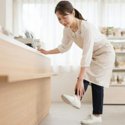 レジに立つ仕事の合間に、足の前側の痛みを気にして靴に手を当てる女性店員(イメージ)